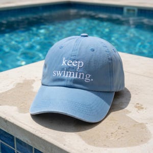 May include: A light blue baseball cap with the embroidered text "keep swimming." The cap has a curved brim and a small button on top. The hat is made of a soft fabric and is perfect for a day at the pool or beach.