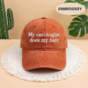 May include: A rust-colored baseball cap with the embroidered text "My oncologist does my hair" in white. The cap is sitting on a white, fringed, circular mat. The word "EMBROIDERY" is in a white oval in the background.
