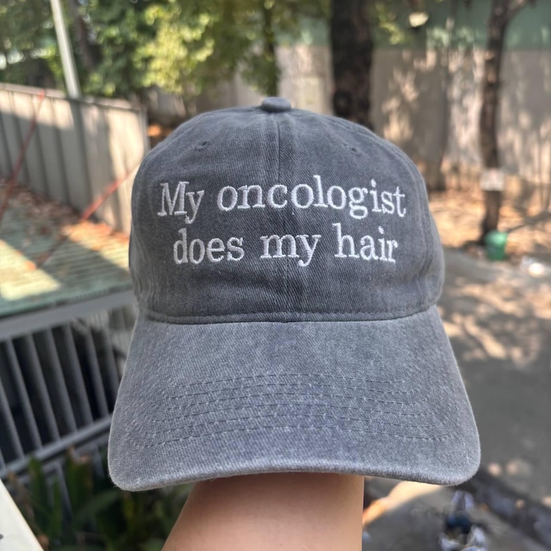 May include: Gray baseball cap with the embroidered text "My oncologist does my hair". The cap has a curved brim and a fabric strap closure. The hat is made of a soft, washed cotton material.