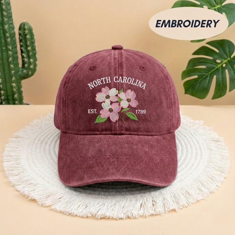 May include: A maroon baseball cap with the embroidered text "NORTH CAROLINA" and "EST. 1789". The cap features a floral design with pink and green accents. The hat is sitting on a white, fringed surface.