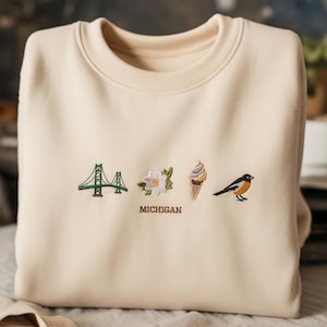 May include: Cream-colored sweatshirt with embroidered Michigan-themed designs. Features a green bridge, white flower, ice cream cone, and a bird. The word "MICHIGAN" is embroidered below the designs.