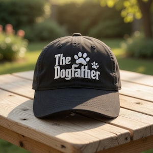May include: Black baseball cap with the text "The Dogfather" in white, along with a paw print design. The cap is sitting on a wooden table outdoors.