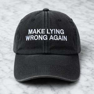 May include: A black baseball cap with the phrase "MAKE LYING WRONG AGAIN" embroidered in white. The cap has a curved brim and a small button on top. The hat is made of a soft, worn material.