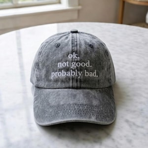 May include: A gray baseball cap with the text "ok. not good. probably bad." embroidered in white. The cap has a curved brim and a button on top. The hat is made of a washed, textured fabric.