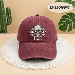 Puede incluir: Una gorra de b&eacute;isbol color granate con acabado desgastado que presenta un bordado blanco de una calavera con tibias cruzadas. El dise&ntilde;o incluye los n&uacute;meros 86 y 47 a ambos lados de la calavera, con las letras FDT impresas en blanco debajo.