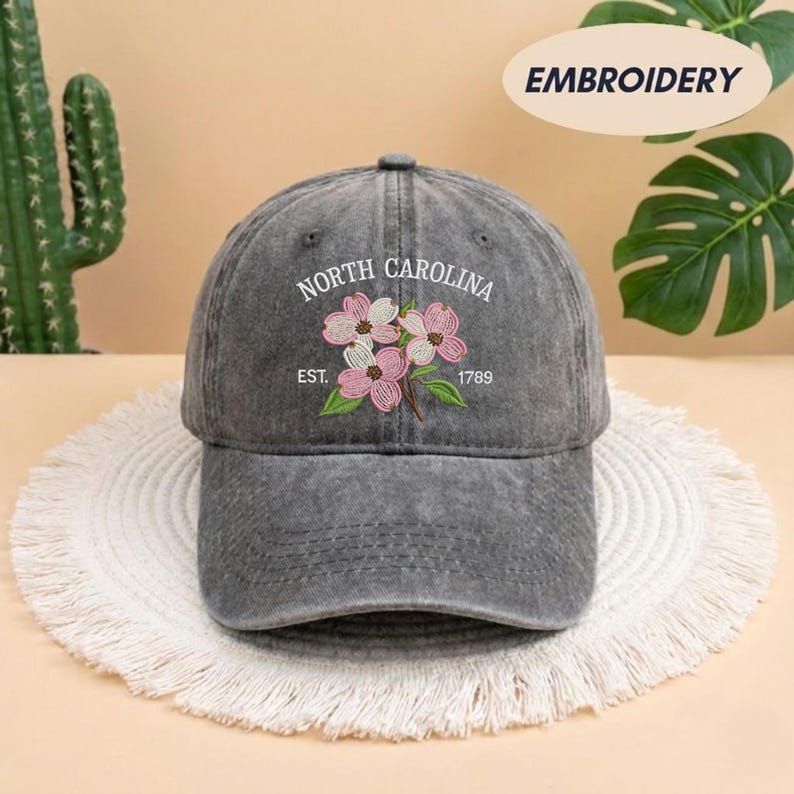 May include: A gray baseball cap with the embroidered text "NORTH CAROLINA" and "EST. 1789". The cap features a floral design with pink and white flowers and green leaves. The word "EMBROIDERY" is printed on the top right.