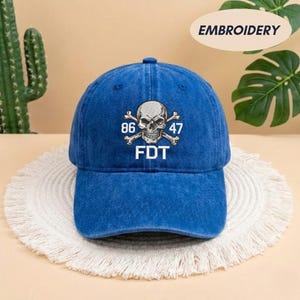 Puede incluir: Una gorra de b&eacute;isbol azul con acabado desgastado que presenta un gr&aacute;fico bordado en blanco de una calavera con tibias cruzadas. El dise&ntilde;o incluye los n&uacute;meros 86 y 47 a ambos lados de la calavera, con las letras FDT impresas en blanco debajo.