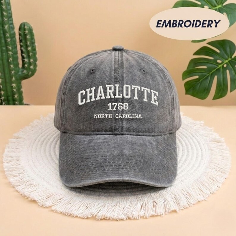May include: A gray baseball cap with the word "CHARLOTTE" in white block letters. Below the word "CHARLOTTE" is the number "1768" and the words "NORTH CAROLINA". The cap is sitting on a white, fringed placemat.