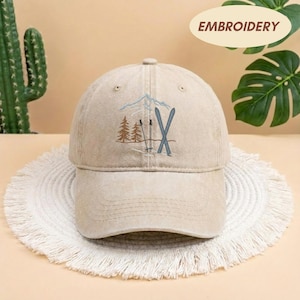 May include: A beige baseball cap with an embroidered design featuring a mountain, skis, and trees. The word "EMBROIDERY" is printed on a cream-colored oval. The cap is sitting on a white, fringed surface.