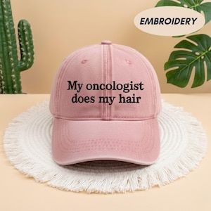 May include: A dusty rose baseball cap with the embroidered text "My oncologist does my hair" in black. The cap is sitting on a white, fringed mat. The word "EMBROIDERY" is in a white oval in the background.