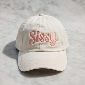 May include: A cream-colored baseball cap with the word "Sissy" embroidered in pink script across the front. The cap has a curved brim and a button on top.