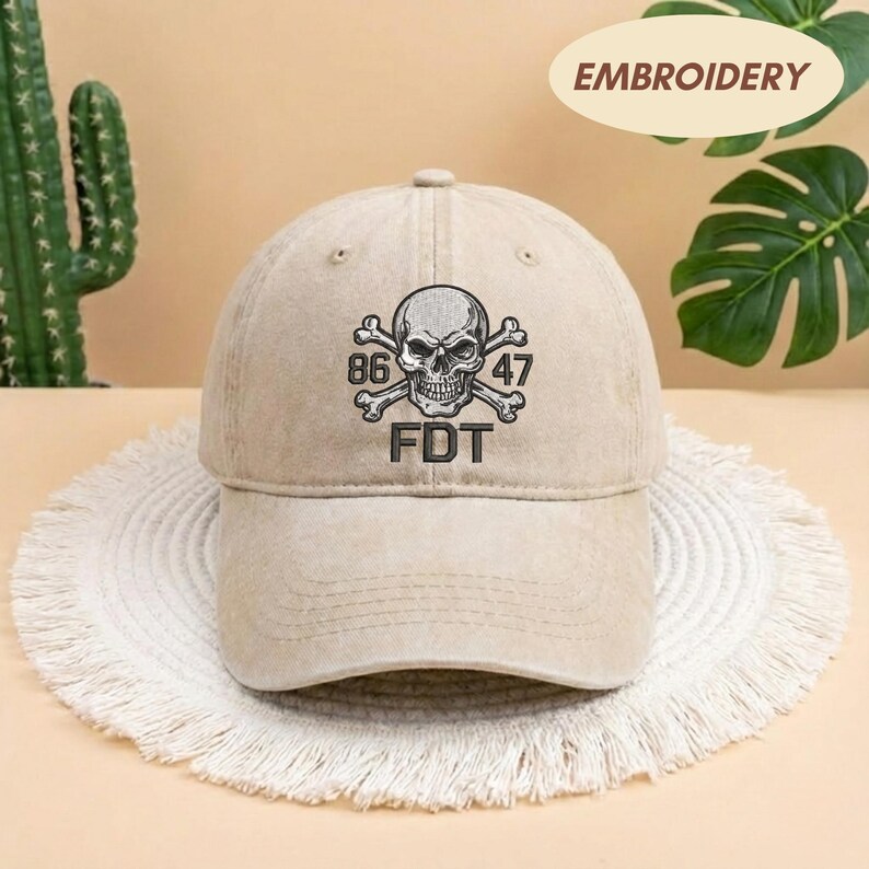 Puede incluir: Una gorra de b&eacute;isbol de color beige con un dise&ntilde;o bordado de una calavera y tibias cruzadas, con los n&uacute;meros 86 y 47 a ambos lados. Debajo de la calavera, las letras FDT est&aacute;n bordadas en hilo oscuro. La gorra tiene una corona estructurada.