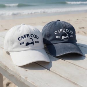 May include: Two baseball caps with the text "CAPE COD EST 1602" in navy blue. One cap is white, the other is navy blue. The caps are sitting on a wooden surface with a beach and ocean in the background.