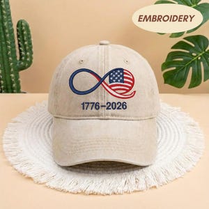 May include: A beige baseball cap with an embroidered design featuring the American flag in a stylized infinity symbol. The design includes the dates 1776-2026 in navy blue. The word "EMBROIDERY" is printed on the top right.