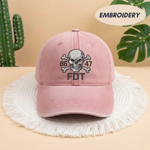 Puede incluir: Una gorra de b&eacute;isbol de color rosa empolvado con un bordado que muestra una calavera y tibias cruzadas. Los n&uacute;meros 86 y 47 est&aacute;n situados a ambos lados de la calavera, con las letras FDT bordadas en hilo negro justo debajo del dise&ntilde;o.