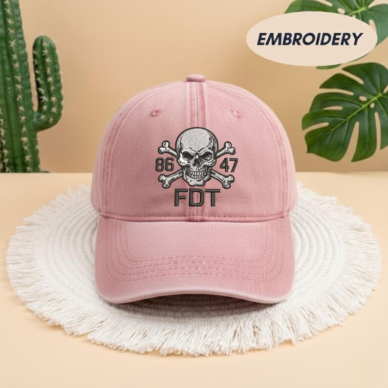 Puede incluir: Una gorra de b&eacute;isbol de color rosa empolvado con un bordado que muestra una calavera y tibias cruzadas. Los n&uacute;meros 86 y 47 est&aacute;n situados a ambos lados de la calavera, con las letras FDT bordadas en hilo negro justo debajo del dise&ntilde;o.
