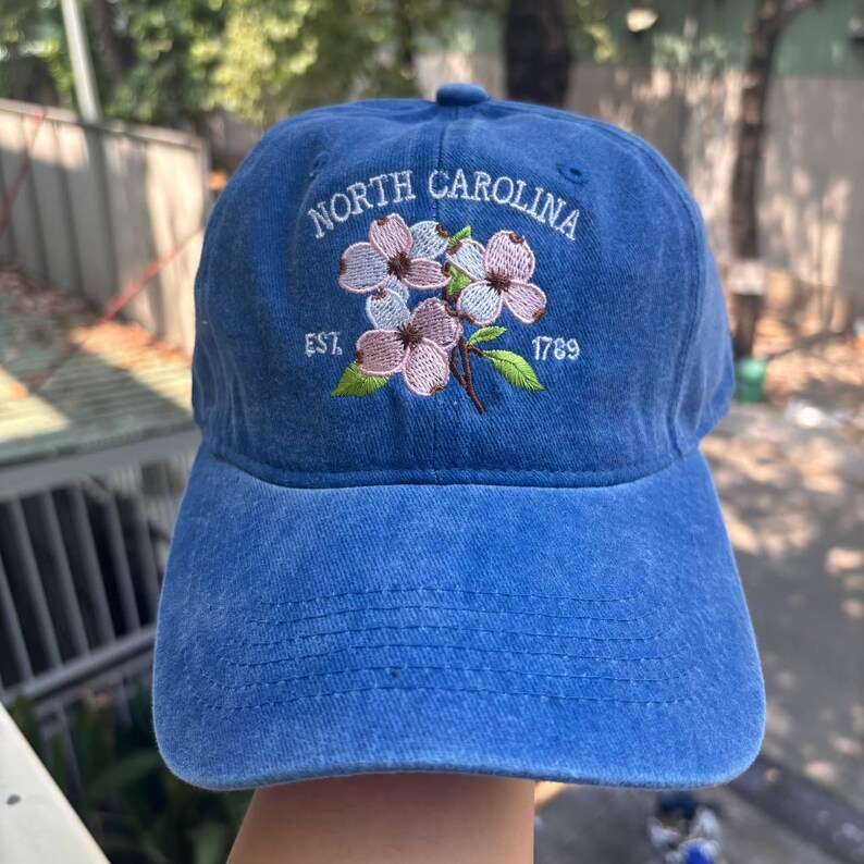 May include: Blue baseball cap with embroidered floral design and text "North Carolina EST. 1789". The cap features a curved brim and adjustable strap. The embroidery includes pink and white flowers with green leaves.