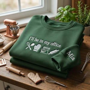 May include: A forest green crewneck sweatshirt featuring white embroidery that reads "I'll be in my office" above icons of a whisk, oven mitt, stand mixer, and rolling pin. The name "Mia" is embroidered in white on the sleeve cuff.