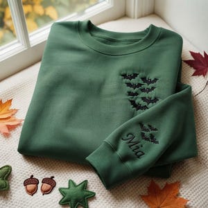 May include: A forest green crewneck sweatshirt featuring black embroidered bats on the chest and a sleeve cuff. The sleeve cuff also displays the name Mia embroidered in black script lettering, accompanied by a small cluster of black embroidered bats.