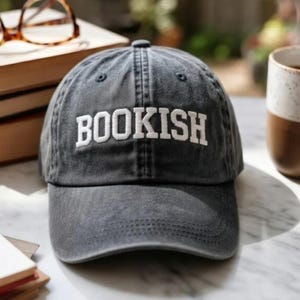 May include: A gray baseball cap with the word "BOOKISH" embroidered in white. The hat has a curved brim and a classic six-panel design. The cap is made of a soft, worn fabric.