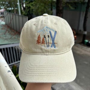 May include: A beige baseball cap with an embroidered design featuring a mountain, skis, and trees in shades of blue, orange, and brown. The cap has a curved brim and a button on top, suitable for outdoor activities.