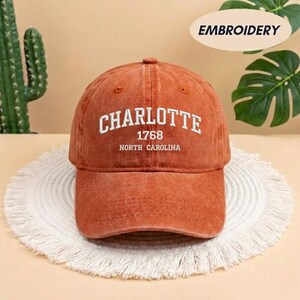 May include: A burnt orange baseball cap with the word "CHARLOTTE" embroidered in white, along with "1768" and "NORTH CAROLINA". The cap is sitting on a white, fringed mat.