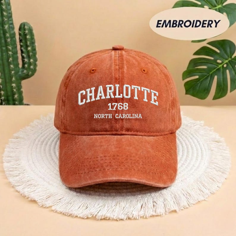 May include: A burnt orange baseball cap with the word "CHARLOTTE" embroidered in white, along with "1768" and "NORTH CAROLINA". The cap is sitting on a white, fringed mat.