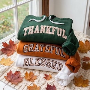 May include: A stack of three hooded sweatshirts in dark green, orange, and white. The green hoodie features the word THANKFUL, the orange hoodie features GRATEFUL, and the white hoodie features BLESSED, all in a plaid-patterned block letter font.