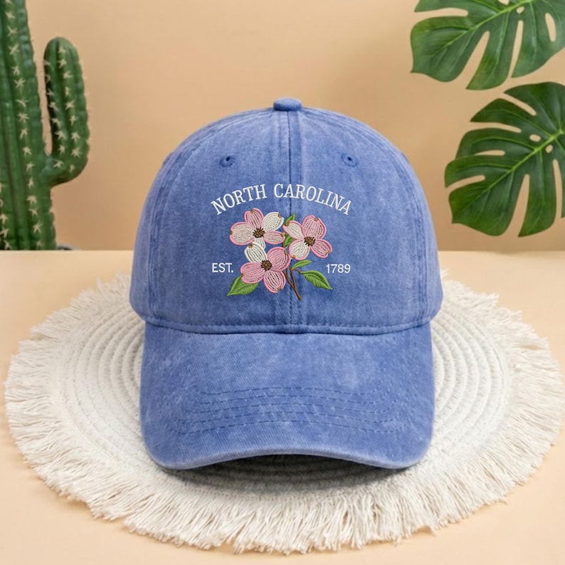 May include: A blue baseball cap with the words "North Carolina" and "Est. 1789" embroidered in white. The cap features an embroidered floral design with pink and white flowers and green leaves. The hat is sitting on a white, fringed surface.
