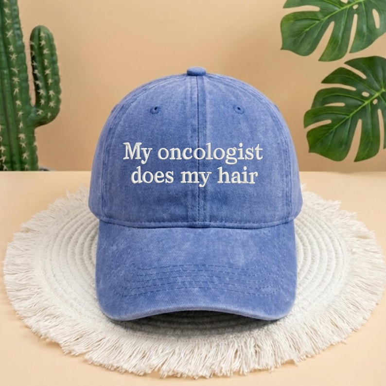 May include: A blue baseball cap with the text "My oncologist does my hair" in white. The cap is made of a soft material and has a curved brim. The hat is sitting on a white, fringed mat.