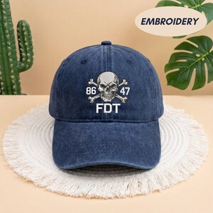 Puede incluir: Una gorra de b&eacute;isbol de mezclilla azul oscuro con un dise&ntilde;o bordado en blanco de una calavera y tibias cruzadas en la parte frontal. El bordado incluye los n&uacute;meros 86 y 47 a ambos lados de la calavera, con las letras FDT en blanco debajo.