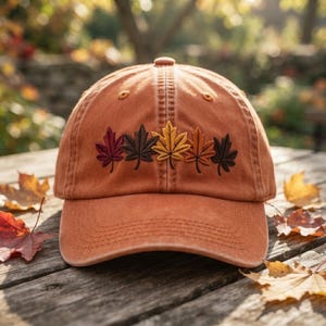May include: A burnt orange baseball cap featuring an embroidered row of five maple leaves in varying autumn shades of deep red, dark brown, golden yellow, and copper orange across the front panels above the curved brim.