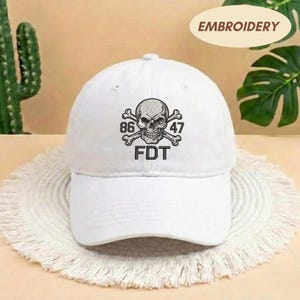 Puede incluir: Una gorra de b&eacute;isbol blanca con un dise&ntilde;o bordado en negro en la parte frontal. El dise&ntilde;o incluye una calavera centrada sobre huesos cruzados, con el n&uacute;mero 86 a la izquierda, 47 a la derecha y las letras FDT impresas en negro debajo.