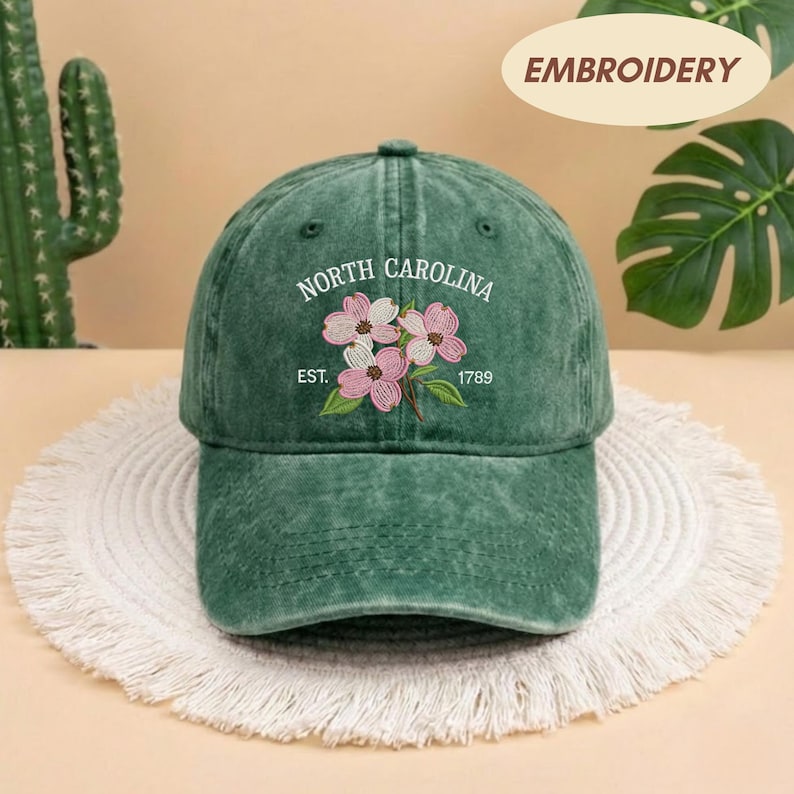 May include: Green baseball cap with embroidered text and floral design. The text reads "North Carolina" above a floral design with the text "Est. 1789". The cap is sitting on a white, circular, woven mat.
