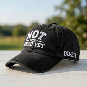 May include: A black baseball cap featuring white embroidered text on the front that reads NOT DEAD YET with a small skull and wings icon between the words. The side of the cap displays the white embroidered text DD-214 on the side panel.