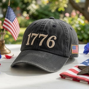 May include: A charcoal grey baseball cap featuring the embroidered text 1776 in a light tan font on the front panel. A small rectangular patch of the United States flag is stitched onto the side of the cap, which has a curved brim and eyelets.