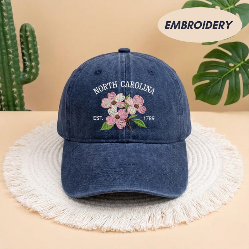 May include: A denim blue baseball cap with embroidered floral design. The cap features the text "NORTH CAROLINA" above a floral design with the text "EST. 1789". The cap is sitting on a white, fringed surface.