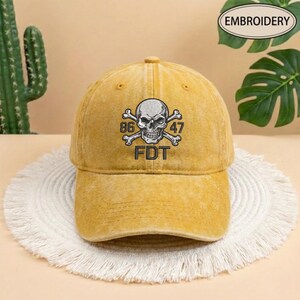 Puede incluir: Una gorra de b&eacute;isbol color amarillo mostaza con acabado desgastado. El frente presenta un dise&ntilde;o bordado de una calavera con huesos cruzados, el n&uacute;mero 86 a la izquierda, 47 a la derecha y las letras FDT centradas bajo la calavera en hilo negro.