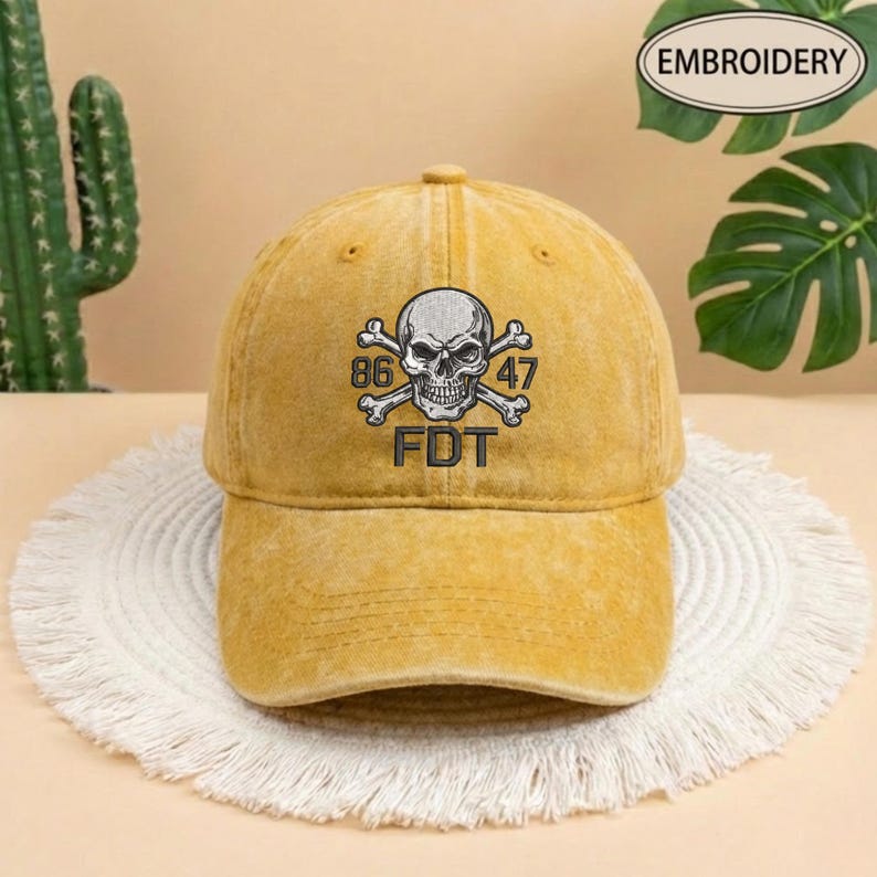 Puede incluir: Una gorra de b&eacute;isbol color amarillo mostaza con acabado desgastado. El frente presenta un dise&ntilde;o bordado de una calavera con huesos cruzados, el n&uacute;mero 86 a la izquierda, 47 a la derecha y las letras FDT centradas bajo la calavera en hilo negro.