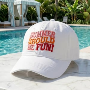 May include: White baseball cap with the embroidered text "SUMMER SHOULD BE FUN!" in shades of pink, orange, and red. The cap has a curved brim and a button on top. The background includes a swimming pool and tropical foliage.