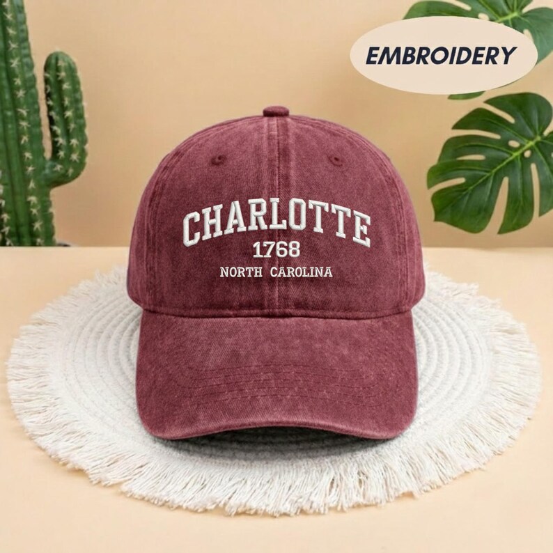 May include: A maroon baseball cap with the word "CHARLOTTE" embroidered in white, along with "1768" and "NORTH CAROLINA". The cap is sitting on a white, fringed mat.