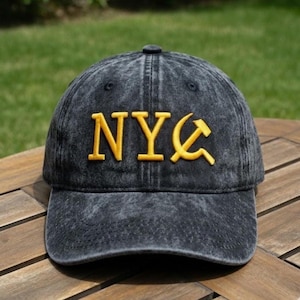 May include: A black, distressed baseball cap with the letters "NYC" embroidered in gold. The "C" is stylized with a hammer and sickle design. The hat is sitting on a wooden surface.