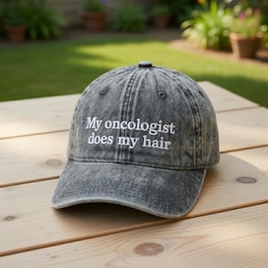 May include: A gray denim baseball cap with the embroidered text "My oncologist does my hair." The hat is sitting on a wooden picnic table outdoors. The hat has a curved brim and a button on top.