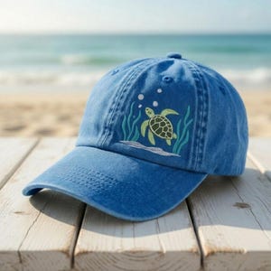 May include: A blue baseball cap with a curved brim. The cap features an embroidered design of a sea turtle swimming among seaweed and bubbles. The background shows a beach and ocean.