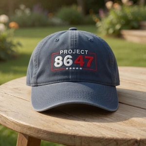 May include: A navy blue baseball cap with the text "PROJECT 8647" in white and red. The cap is sitting on a wooden table outdoors. The hat has a curved brim and a fabric strap for adjustment.