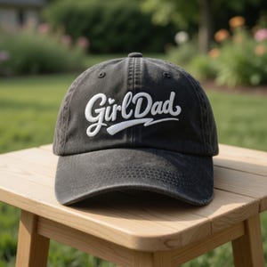 May include: A charcoal gray baseball cap with the embroidered text "Girl Dad" in white script. The hat has a curved brim and is sitting on a wooden table. The background is a blurred outdoor scene.