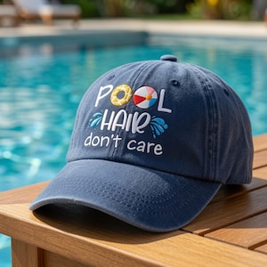 May include: A navy blue baseball cap featuring embroidered text that reads POOL HAIR don't care. The word POOL includes a yellow donut and a multi-colored beach ball as the letter O, with blue water splash graphics surrounding the word HAIR.