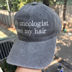 May include: Gray baseball cap with the embroidered text "My oncologist does my hair". The cap has a curved brim and a fabric strap closure. The hat is made of a soft, worn material.