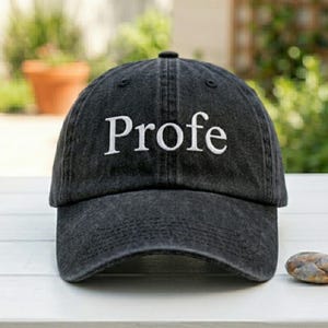 May include: A charcoal gray baseball cap with a washed, distressed texture. The word Profe is embroidered in white, serif-style lettering across the front center of the crown. The cap features a curved brim and a structured six-panel design.