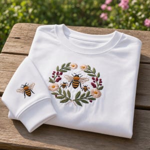 May include: A white long-sleeve sweatshirt featuring an embroidered design of a circular floral wreath with green leaves, pink and white flowers, and three bees in the center. A single bee is also embroidered on the sleeve cuff of the garment.